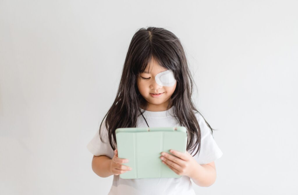 A young girl with a lazy eye and an eye patch on her left eye reads on an iPad.