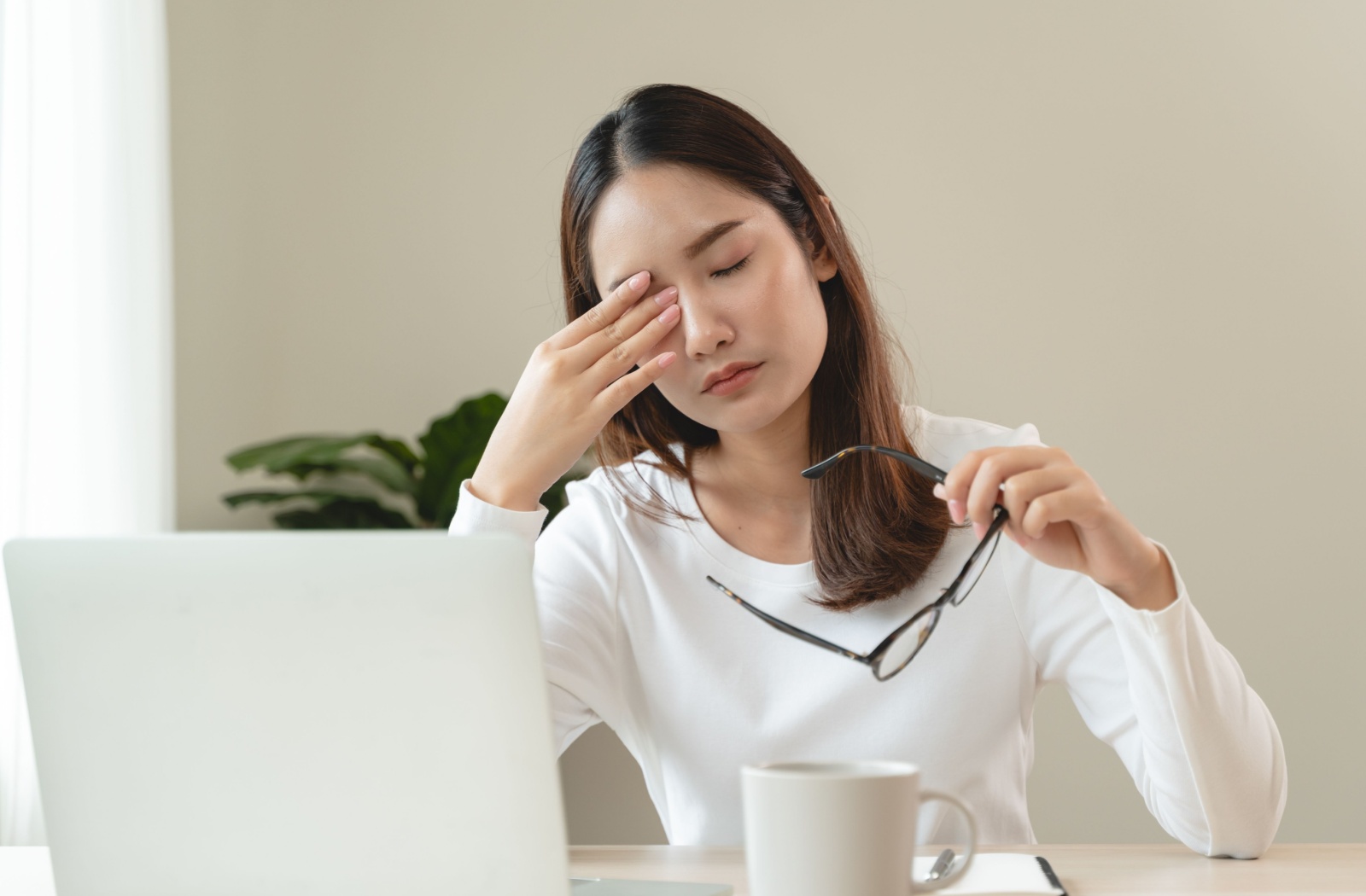 A young adult sitting at a table infront of a laptop rubbing their eye after taking off their glasses.